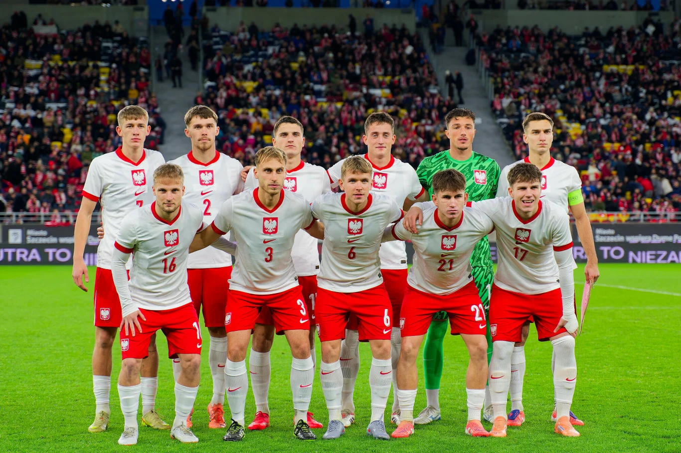 jedenastu młodych piłkarzy w białych koszulkach i czerwonych spodenkach z orłem na piersi stoi w dwóch rzędach na zielonej murawie, przygotowani do meczu narodowej reprezentacji na tle trybun wypełnionych kibicami