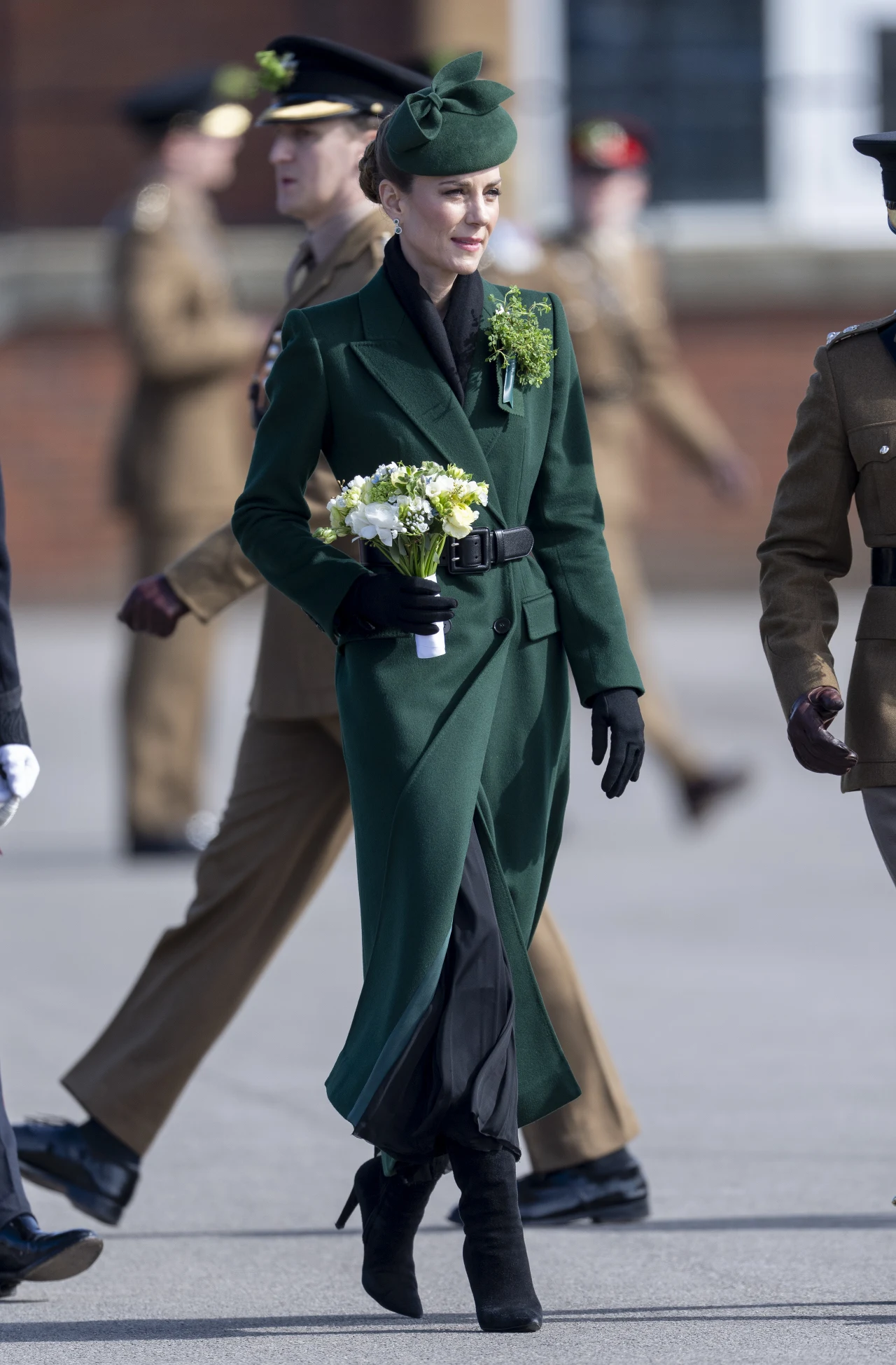 Kobieta w eleganckim, ciemnozielonym płaszczu, czarnych rękawiczkach i kapeluszu w tym samym odcieniu zieleni trzyma bukiet jasnych kwiatów i idzie wśród wojskowych w mundurach, wyróżniając się strojem na tle umundurowanego otoczenia.