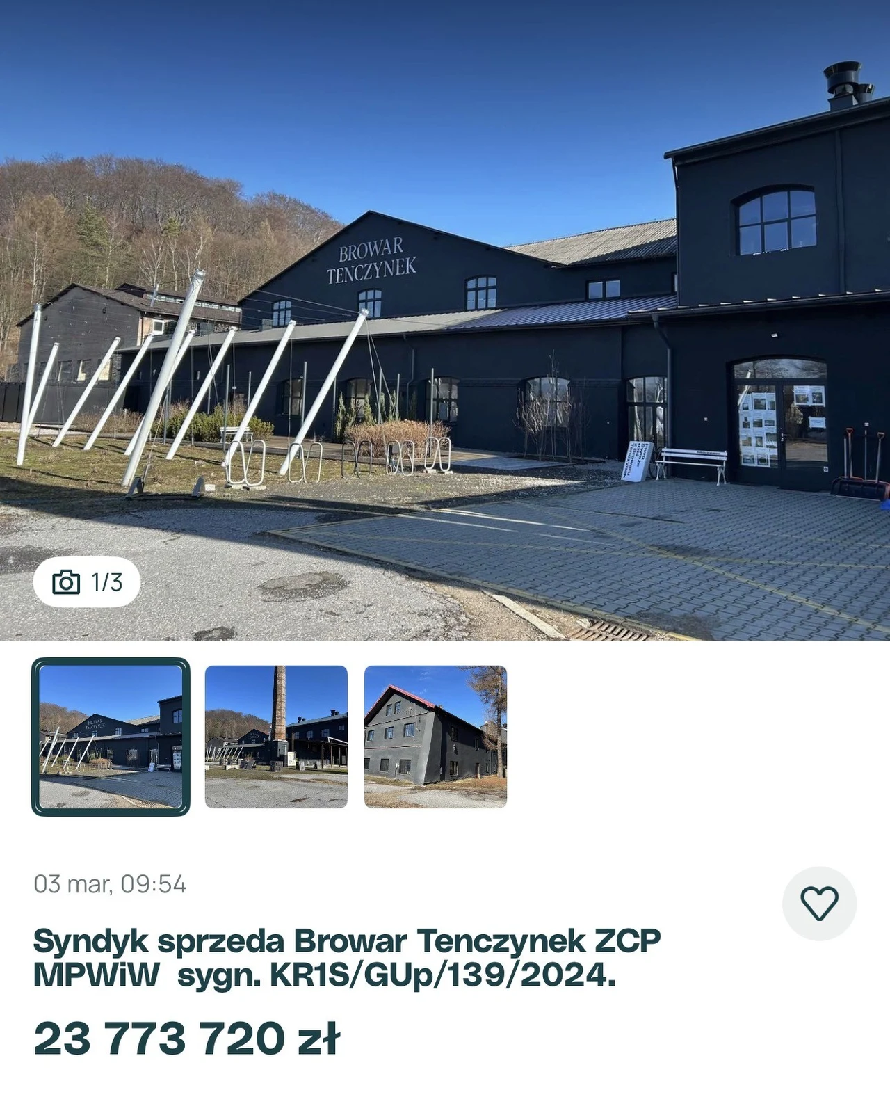 nowoczesny budynek browaru z ciemną elewacją, dużymi oknami i napisem 'Browar Tenczynek' na fasadzie, otoczony utwardzonym placem i metalowymi konstrukcjami, w tle widoczne drzewa i fragment wzgórza