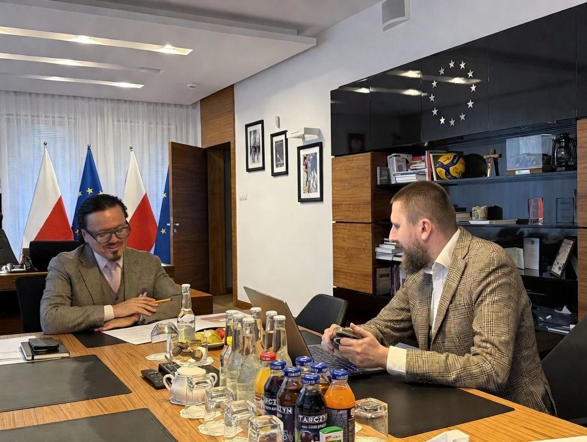 Dwóch mężczyzn w garniturach prowadzi rozmowę przy stole, na którym stoją napoje, filiżanki oraz szklanki; w tle widoczne polskie i unijne flagi oraz półka z książkami i dekoracją w formie gwiazd Europy.