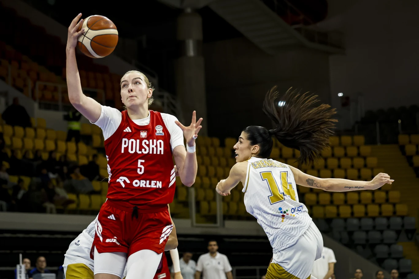 koszykarka reprezentacji Polski w czerwonym stroju z numerem 5 wyciąga rękę po piłkę podczas dynamicznej akcji, obok zawodniczka drużyny przeciwnej w białym stroju z numerem 14 próbuje ją zatrzymać