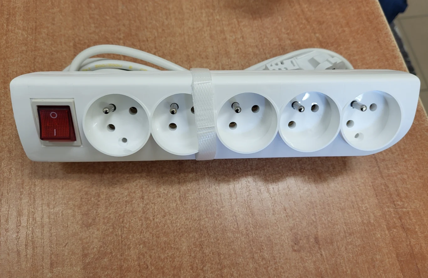 biała listwa zasilająca z pięcioma gniazdami i czerwonym wyłącznikiem, ułożona na drewnianym blacie; przewód zwinięty i spięty opaską