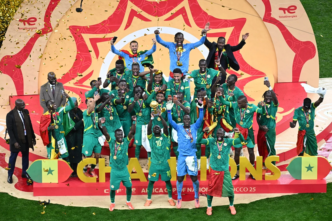 piłkarze drużyny Senegalu w zielonych strojach świętują zdobycie mistrzostwa, na podium unoszą w górę puchar otoczeni medalami i konfetti, za nimi dekoracja z napisem ‚CHAMPIONS’ oraz flagami Senegalu