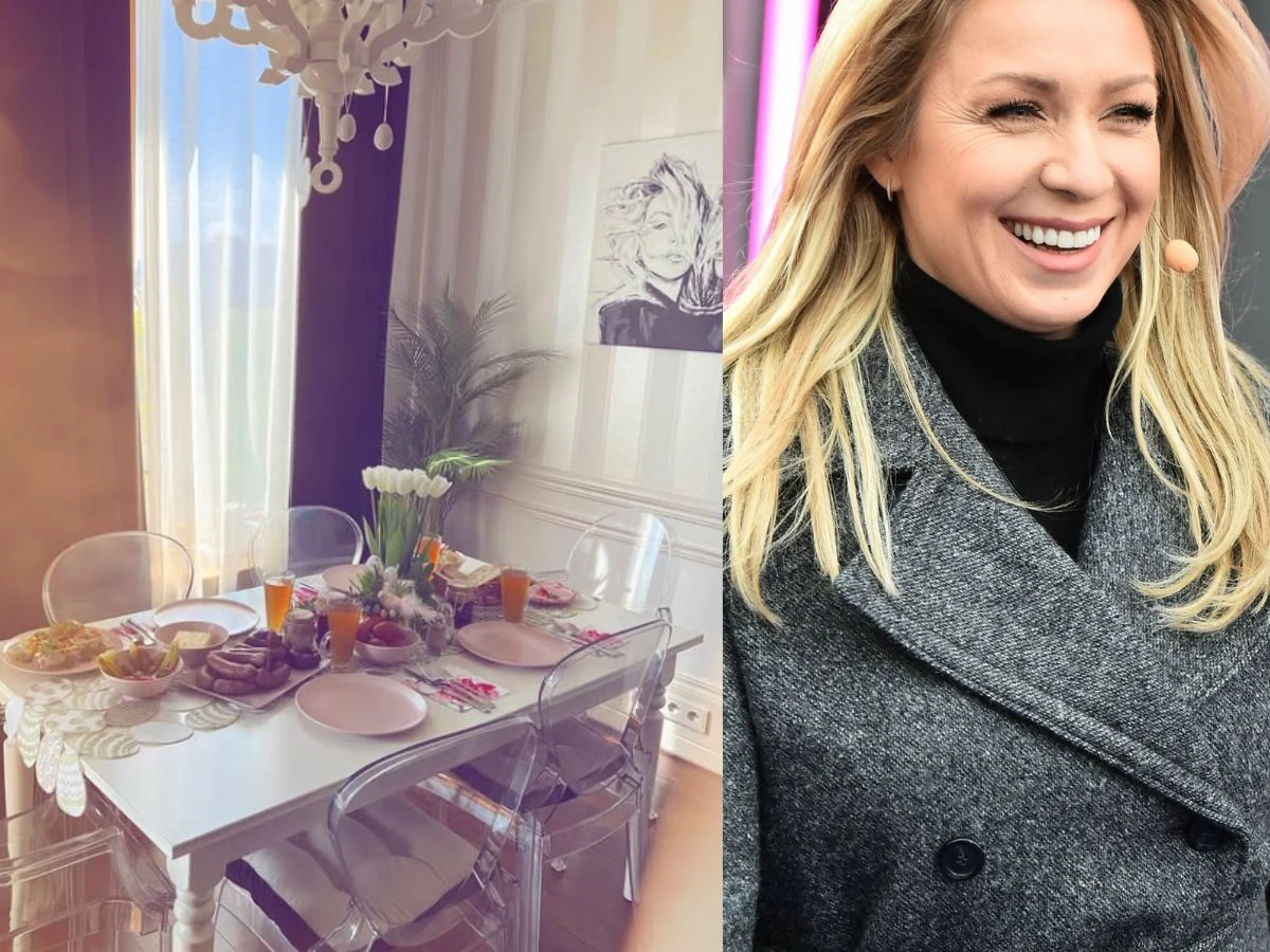 Estetycznie nakryty stół z przezroczystymi krzesłami, białą zastawą i świeżymi kwiatami, na którym stoją półmiski z jedzeniem i napoje, w nowoczesnym, jasnym wnętrzu oraz uśmiechnięta kobieta w szarym płaszczu i czarnym golfie z mikrofonem przy twarzy.