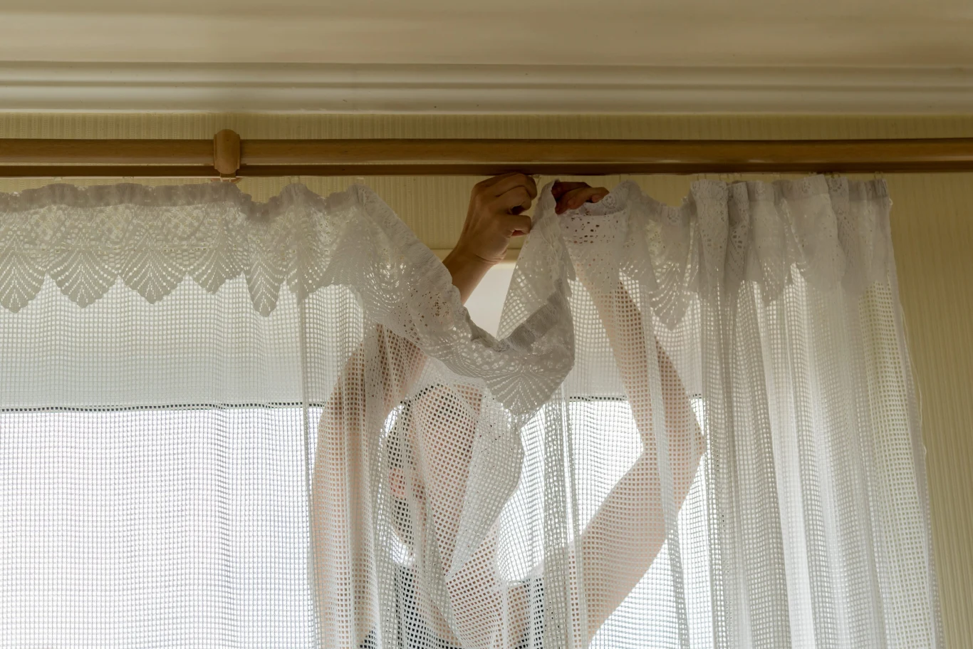 osoba zawiesza koronkową firankę na karniszu, unosząc ręce, by umocować ją na haczykach
