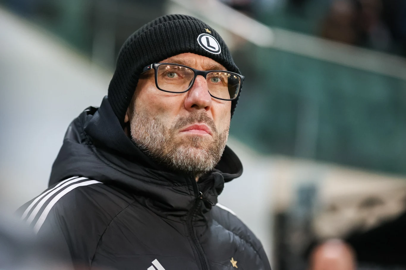 Marek Papszun w okularach i czarnej czapce z herbem Legii na tle rozmytego stadionu