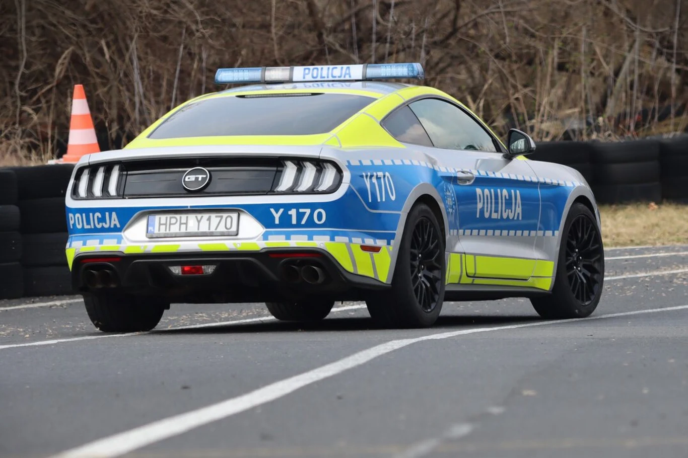 policyjny radiowóz marki Ford Mustang GT w niebiesko-żółtych barwach z widocznym oznaczeniem 'POLICJA', stojący przy drodze asfaltowej, w tle czarne bariery i pomarańczowy pachołek