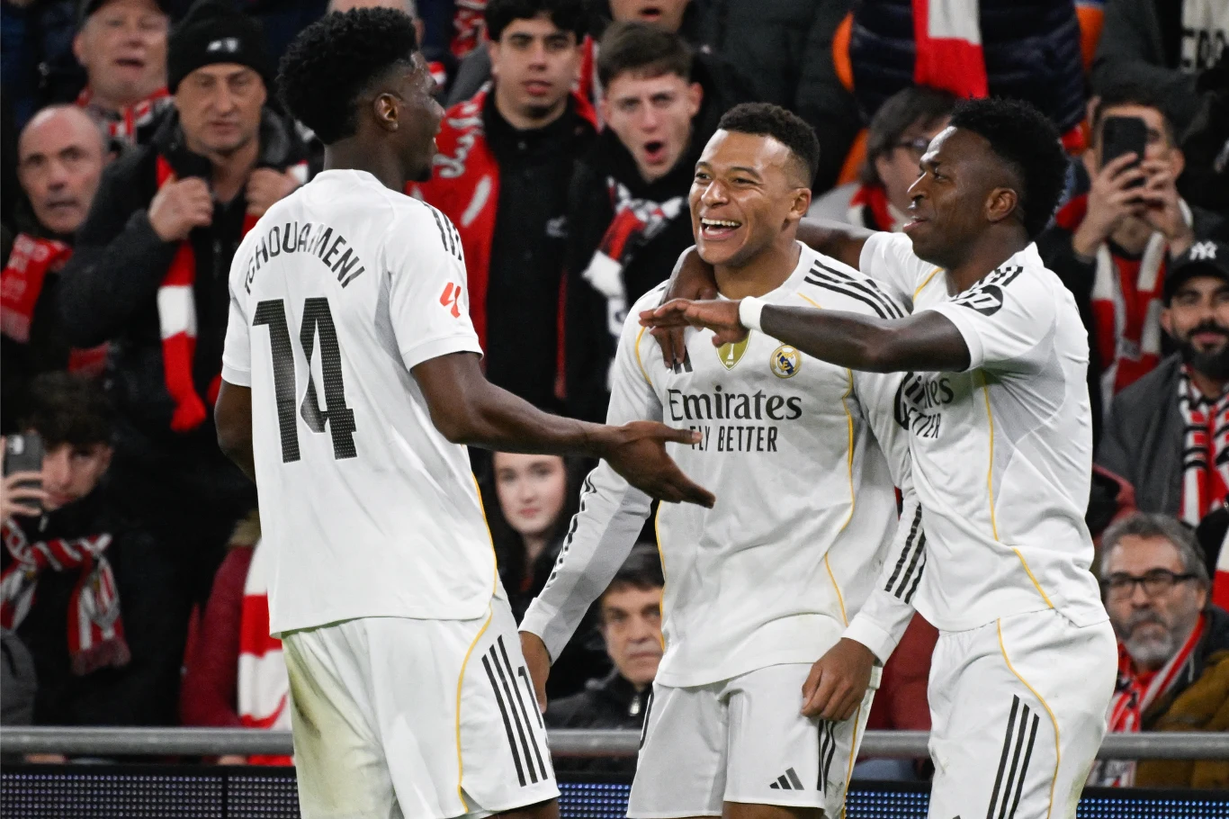 Trzech piłkarzy w białych strojach drużyny Realu Madryt cieszy się po udanej akcji, uśmiechając się i obejmując, podczas gdy w tle widać kibiców na trybunach.