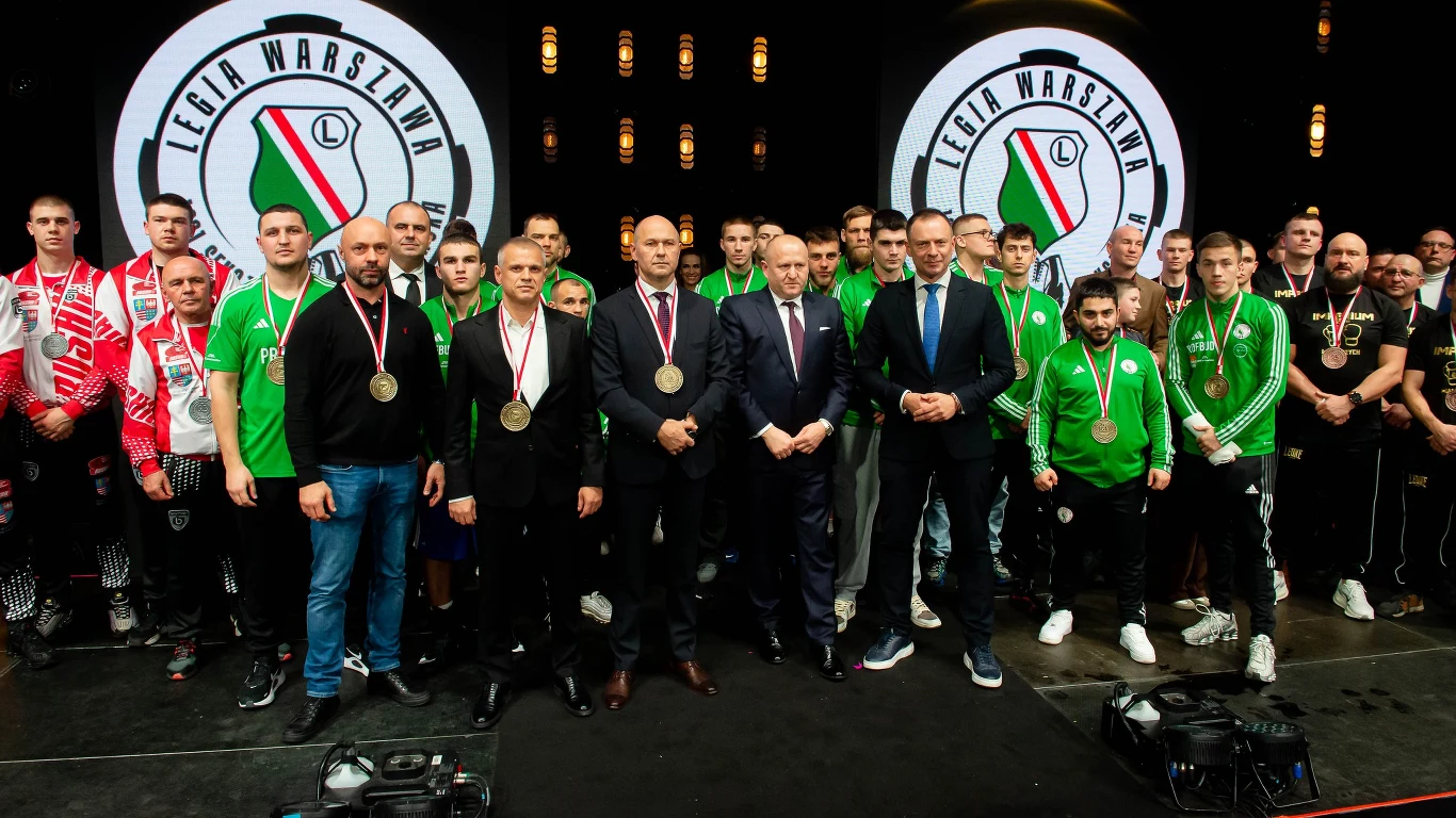 grupa mężczyzn ubranych w sportowe stroje klubowe z medalami na szyjach pozuje razem z kilkoma osobami w garniturach na tle dużych logotypów Legii Warszawa, atmosfera oficjalnego wydarzenia sportowego