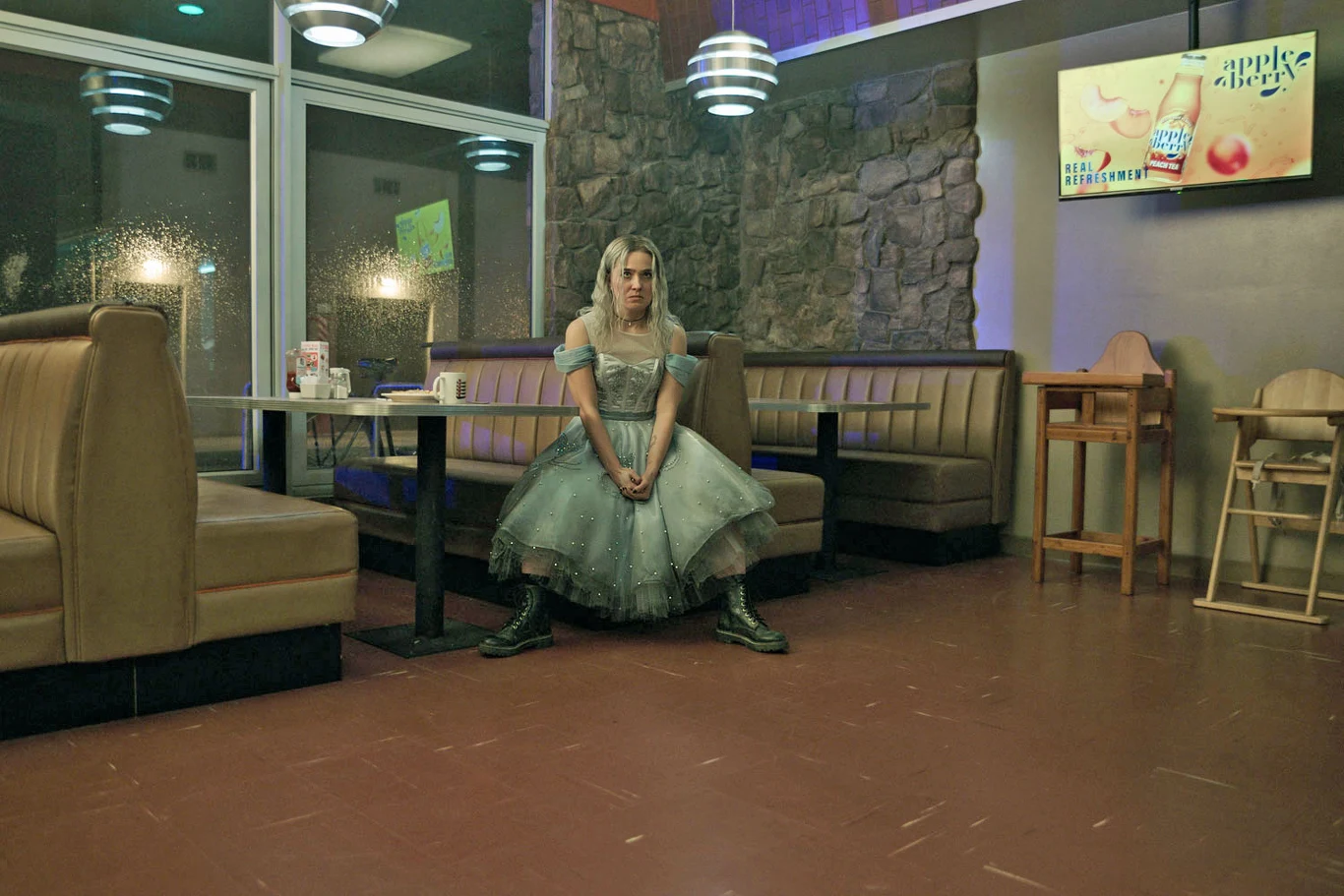 Haley Lu Richardson w "Baw się dobrze i przeżyj" Haley Lu Richardson w sukni ala Alicja z Krainy Czarów siedzi samotnie w pustej kawiarni z nowoczesnym oświetleniem.