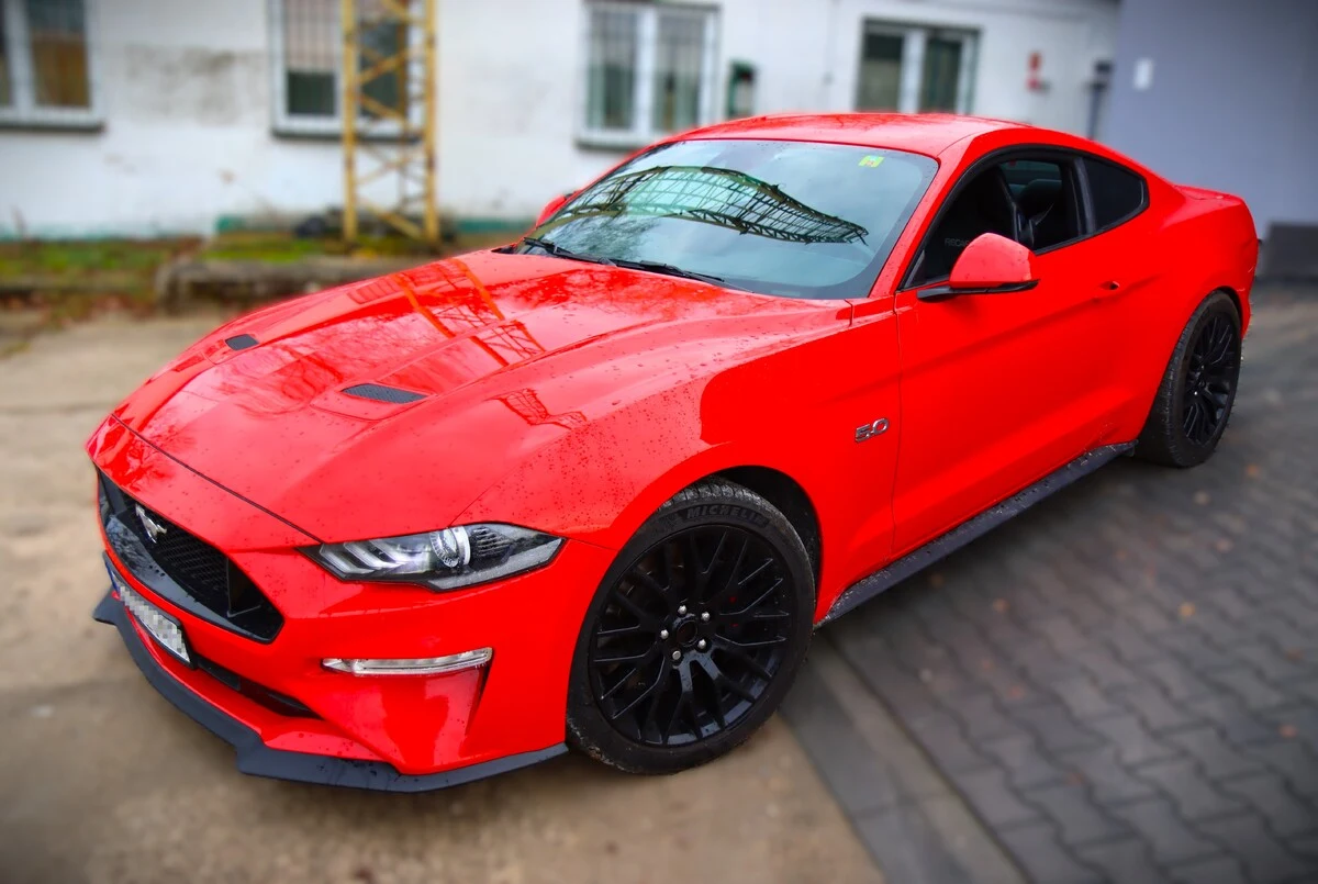 Czerwony sportowy samochód Ford Mustang z czarnymi felgami zaparkowany na chodniku przy budynku z białą elewacją i dużymi oknami.