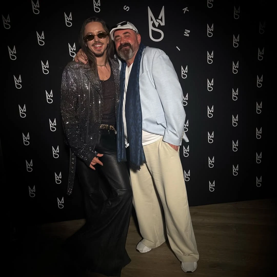 Dwóch mężczyzn stojących razem na tle czarnej ścianki z białymi logotypami; jeden ubrany w błyszczącą kurtkę, ciemne okulary i czarne luźne spodnie, drugi w jasnej marynarce, białej czapce z daszkiem, jasnych spodniach i szaliku, obaj uśmiechnięci, obe...