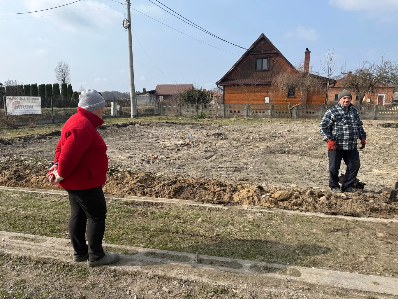 Dwie osoby rozmawiają na tle pustej, przygotowanej działki budowlanej, w oddali drewniany dom z brązowym dachem, otoczony ogrodzeniem i drzewami bez liści.