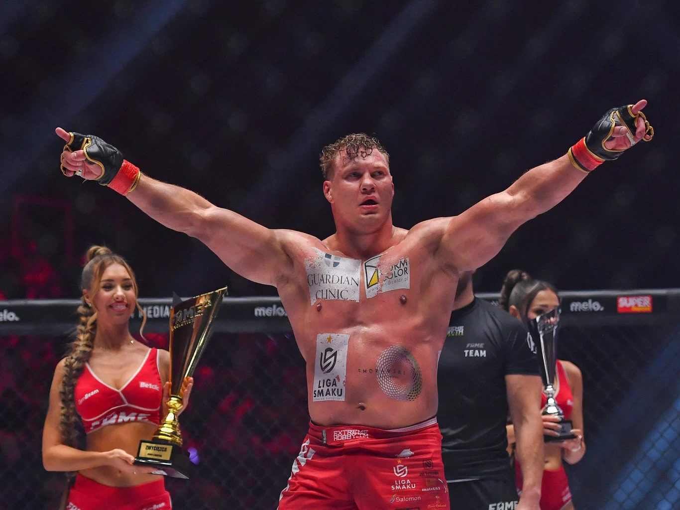 Denis Labryga na ringu z uniesionymi rękami, w tle kobieta z pucharem, sędziowie i trofea