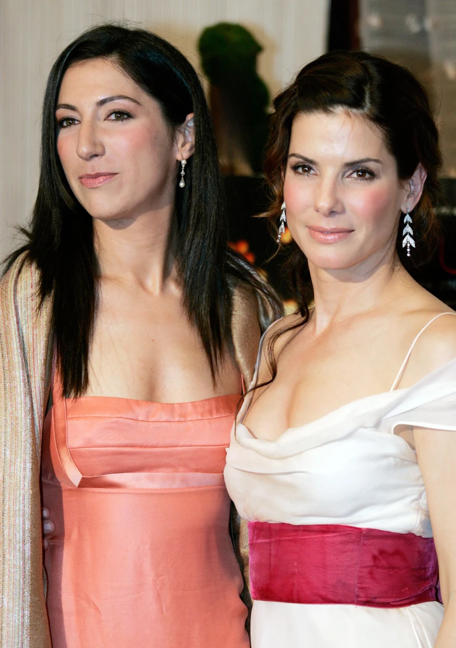 Dwie kobiety pozują razem, obie ubrane elegancko w suknie wieczorowe; jedna w jasnoróżowej kreacji z odkrytymi ramionami i długimi, ciemnymi włosami, druga w białej sukni z szerokim malinowym pasem, nosząca błyszczące kolczyki i z upiętymi włosami.