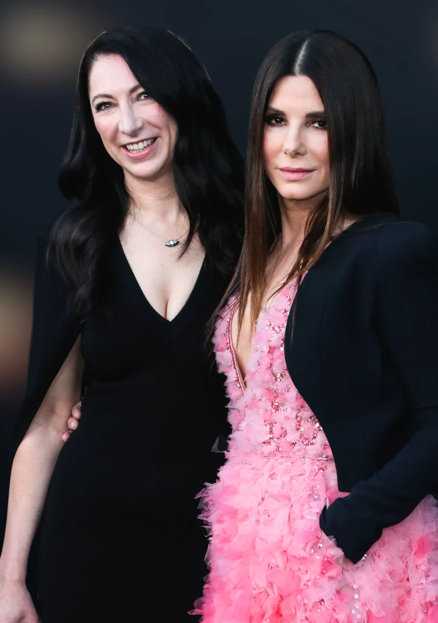 Dwie kobiety stoją obok siebie, jedna ubrana w elegancką, czarną sukienkę z głębokim dekoltem, druga w jasnoróżową suknię ozdobioną piórami z czarną marynarką narzuconą na ramiona; obie uśmiechnięte, z długimi, rozpuszczonymi włosami, patrzą w stronę o...