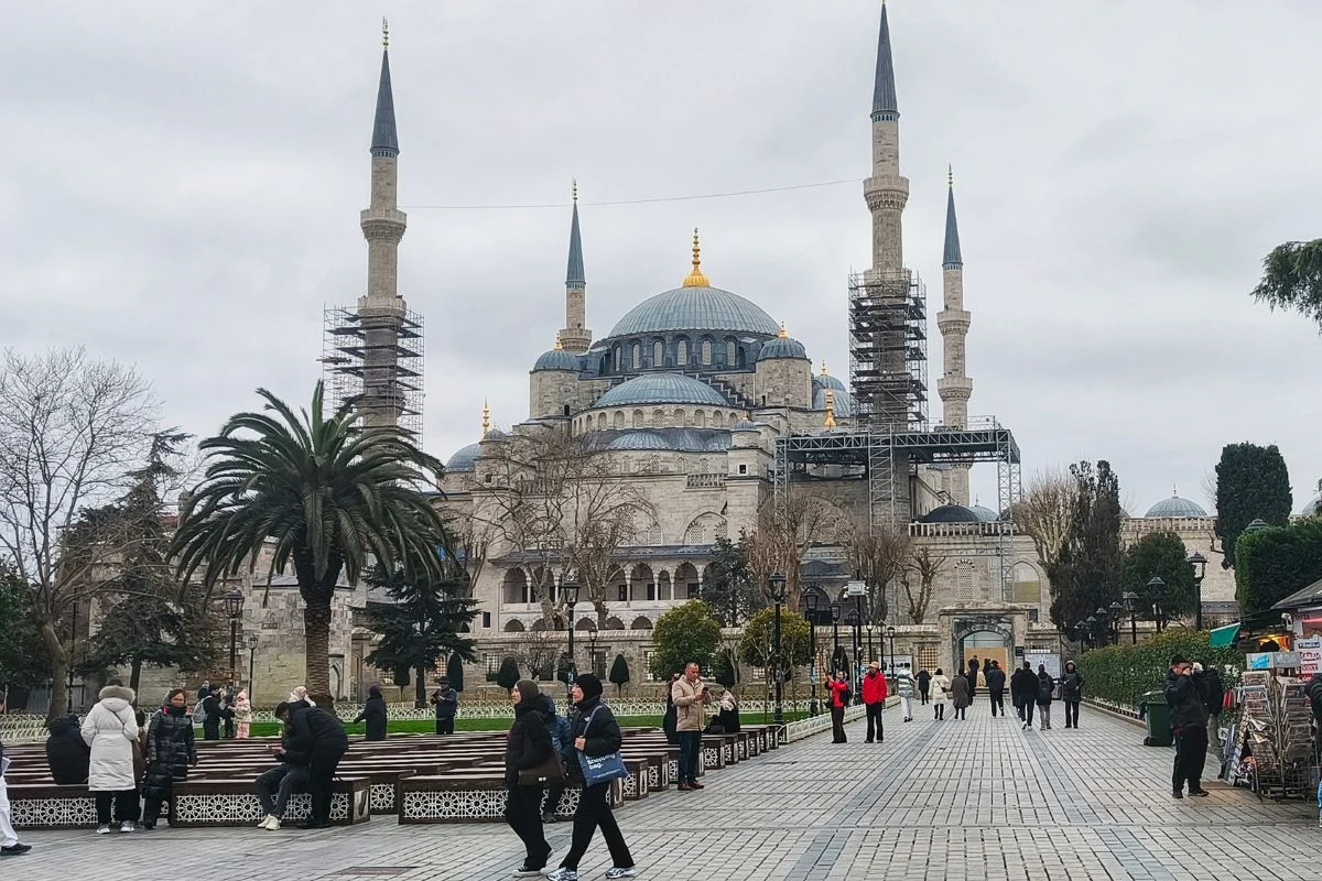 Wielka zabytkowa świątynia z sześcioma smukłymi minaretami i kopułami, wokół której spacerują ludzie; część minaretów otoczona jest rusztowaniami, a przed budowlą rosną palmy i drzewa, tworząc parkowy krajobraz.