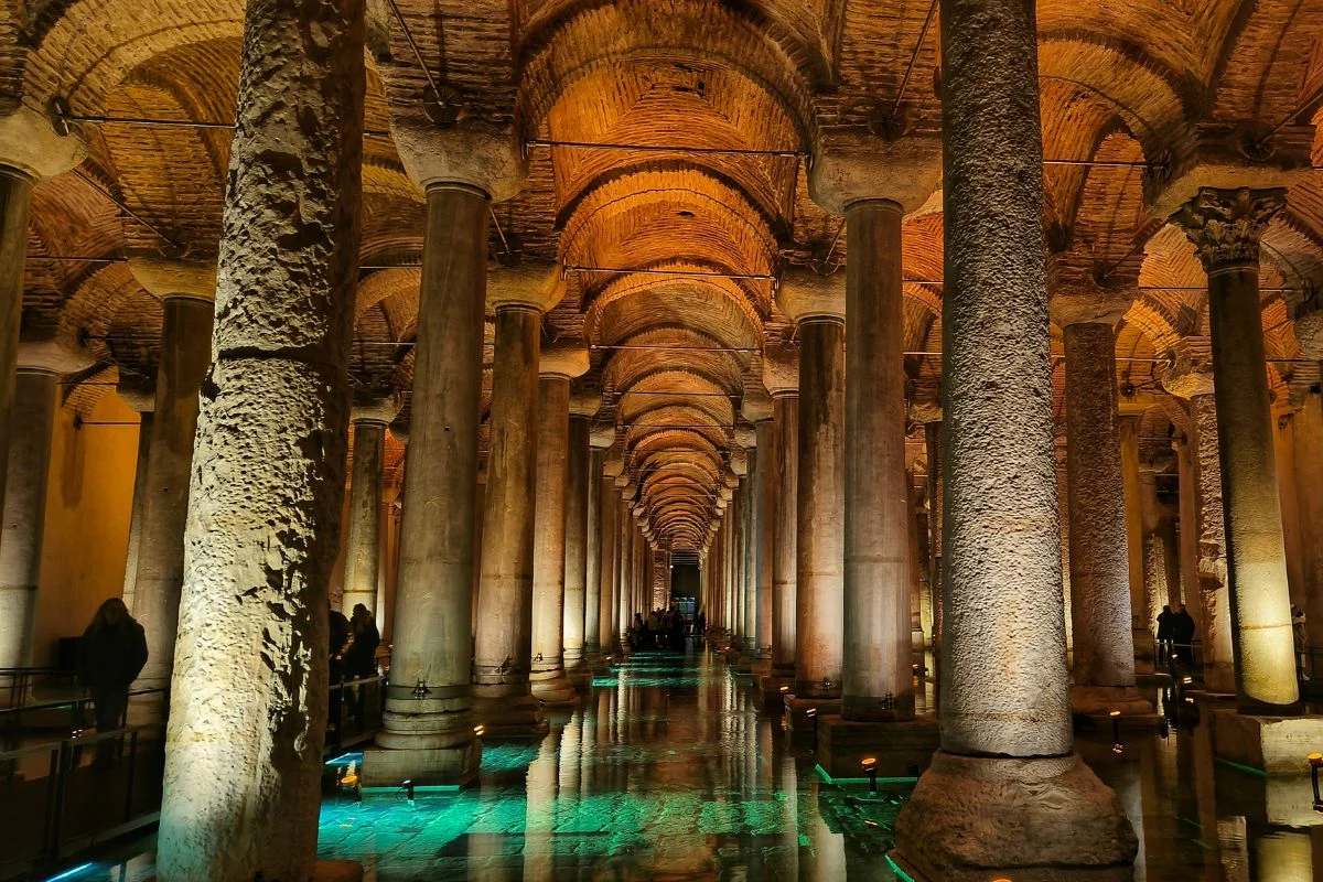 Kamienne kolumny odbijające się w wodzie, sklepienia łukowe oświetlone ciepłym światłem, tworzące rytmiczny, monumentalny ciąg w podziemnej konstrukcji.