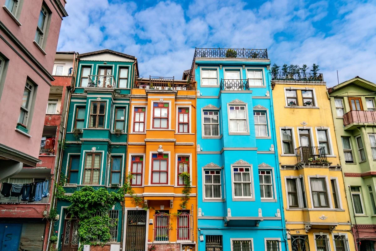 Szereg kolorowych kamienic o ozdobnych fasadach, wśród których wyróżniają się intensywne kolory żółty, niebieski i pomarańczowy, z balkonami, roślinnością pnącą się po jednej ze ścian i jasnym, lekko zachmurzonym niebem w tle.