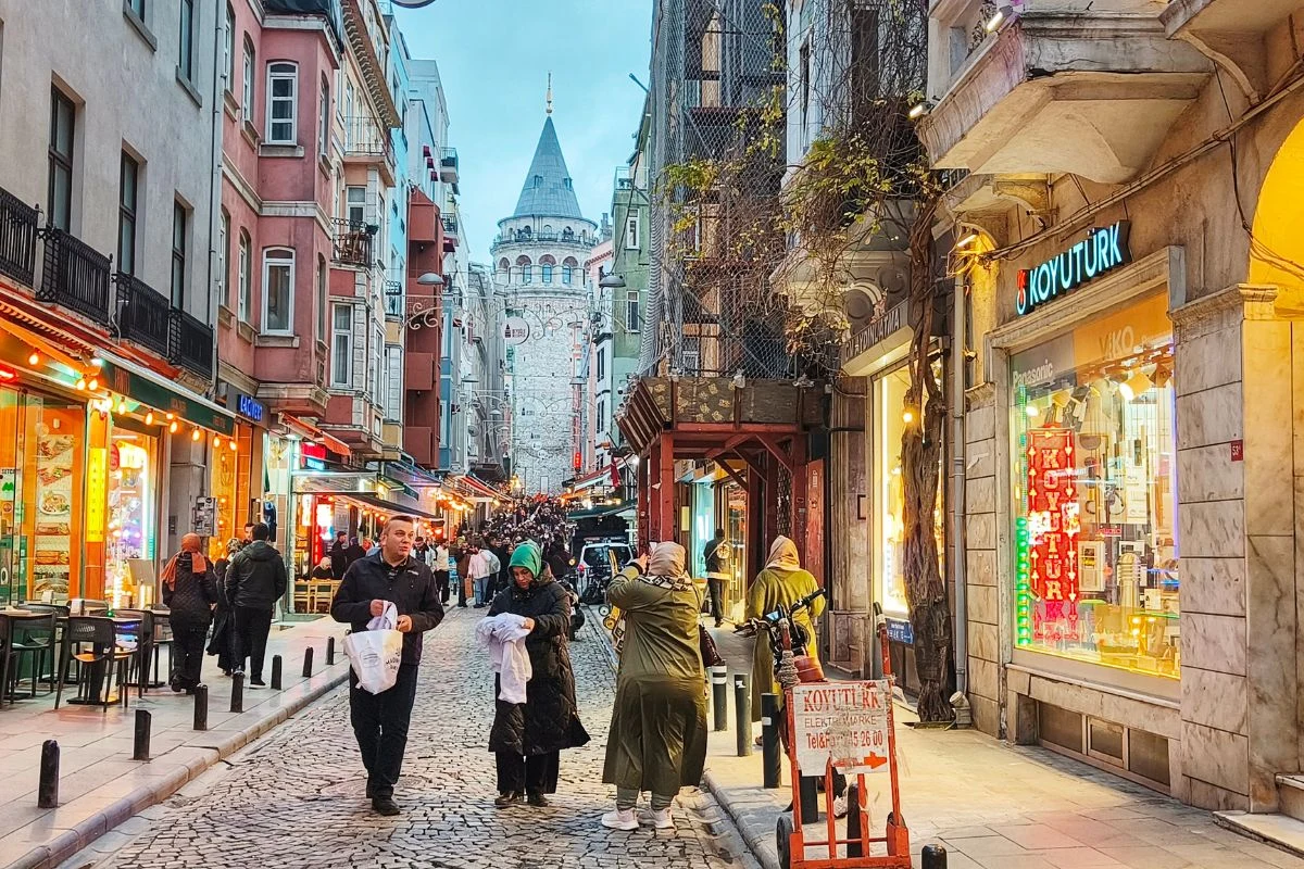 Kolorowa, tętniąca życiem ulica z licznymi kawiarniami i sklepami po bokach, brukowaną nawierzchnią, spacerującymi ludźmi oraz dominującą w tle wieżą Galata.