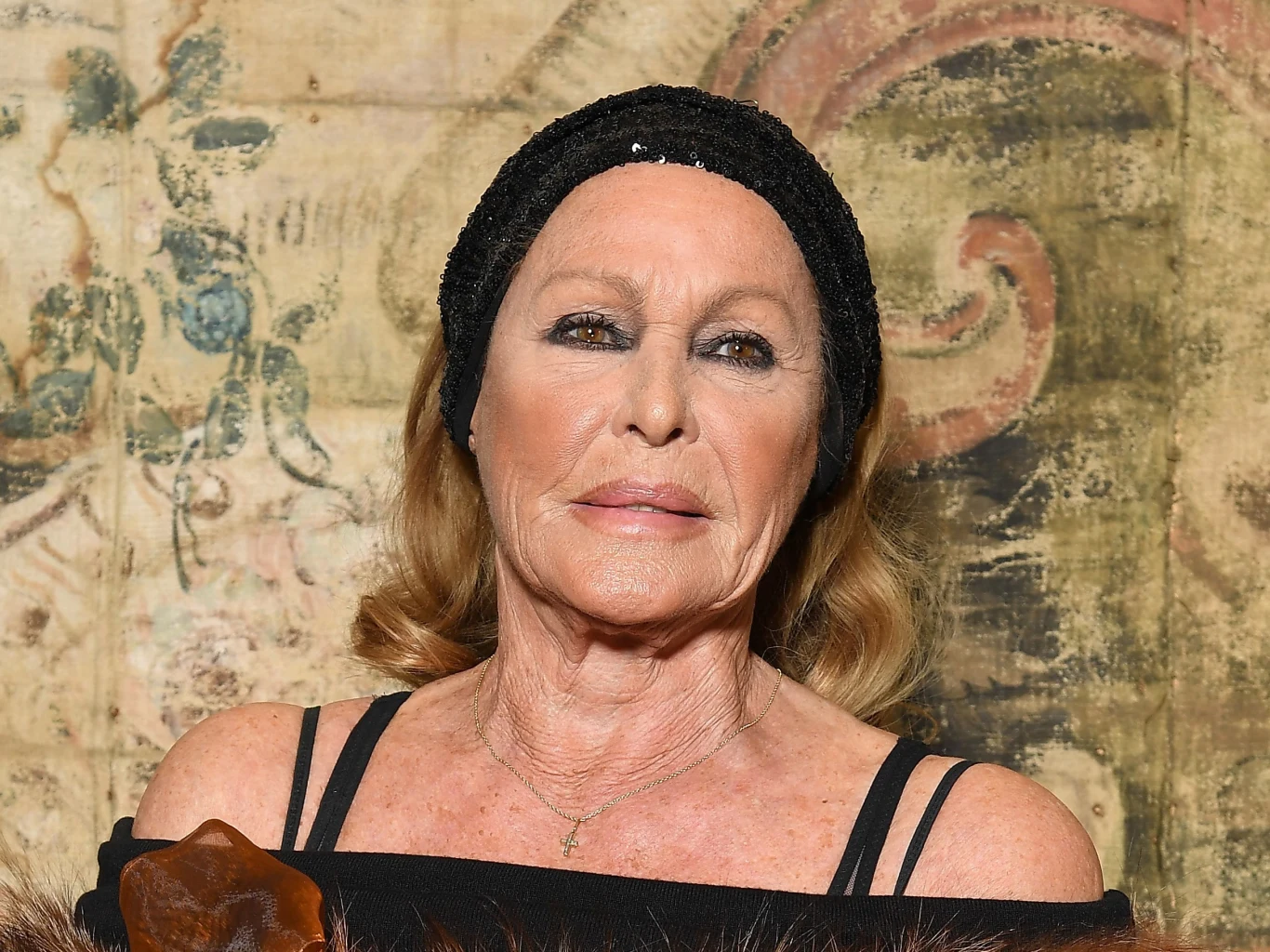 Starsza kobieta z delikatnym makijażem i rozpuszczonymi włosami, ubrana w czarną opaskę oraz czarno-brązowy strój na ramiączkach, patrzy prosto w obiektyw na tle ozdobnej, malowanej ściany o ciepłych barwach.