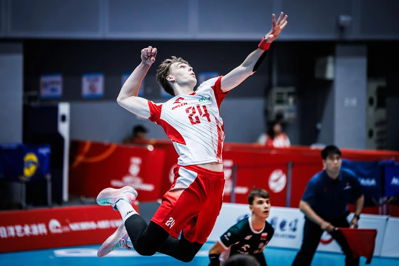 siatkarz w biało-czerwonym stroju, wyskakujący do ataku z dynamicznie wyciągniętą ręką, skupieni zawodnicy i sędzia w tle na arenie sportowej