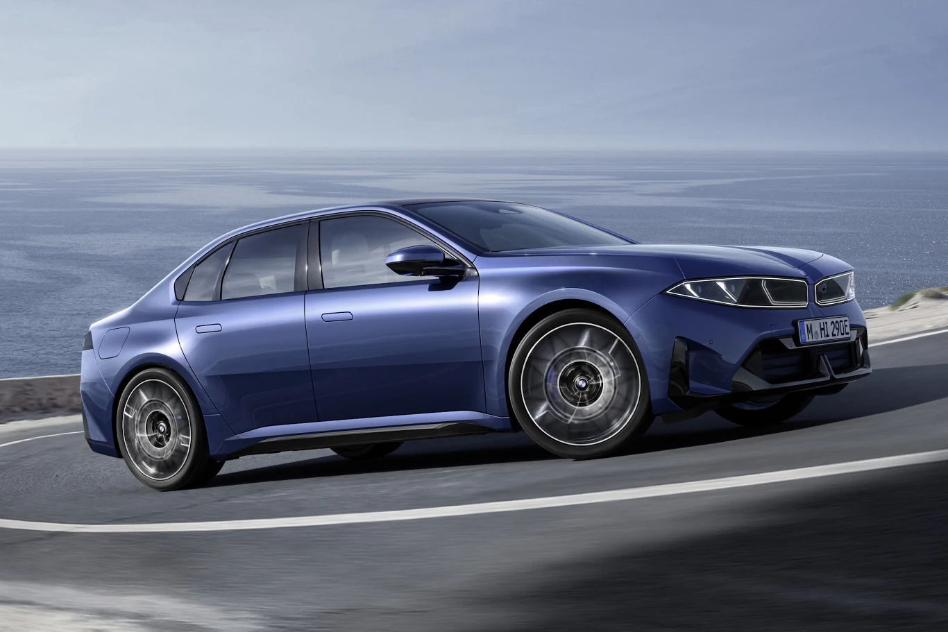 niebieski, sportowy sedan BMW poruszający się dynamicznie po krętej, asfaltowej drodze wzdłuż wybrzeża z widocznym morzem w tle