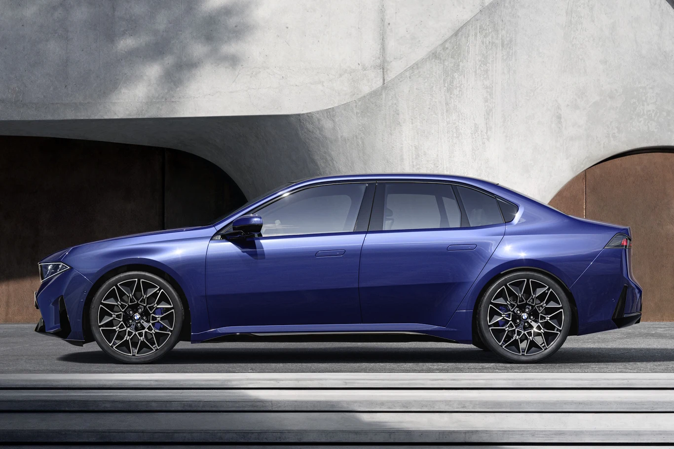 nowoczesny sedan w kolorze niebieskim, o muskularnych liniach nadwozia, dużych aluminiowych felgach i przyciemnianych szybach, zaparkowany na tle minimalistycznej, betonowej architektury