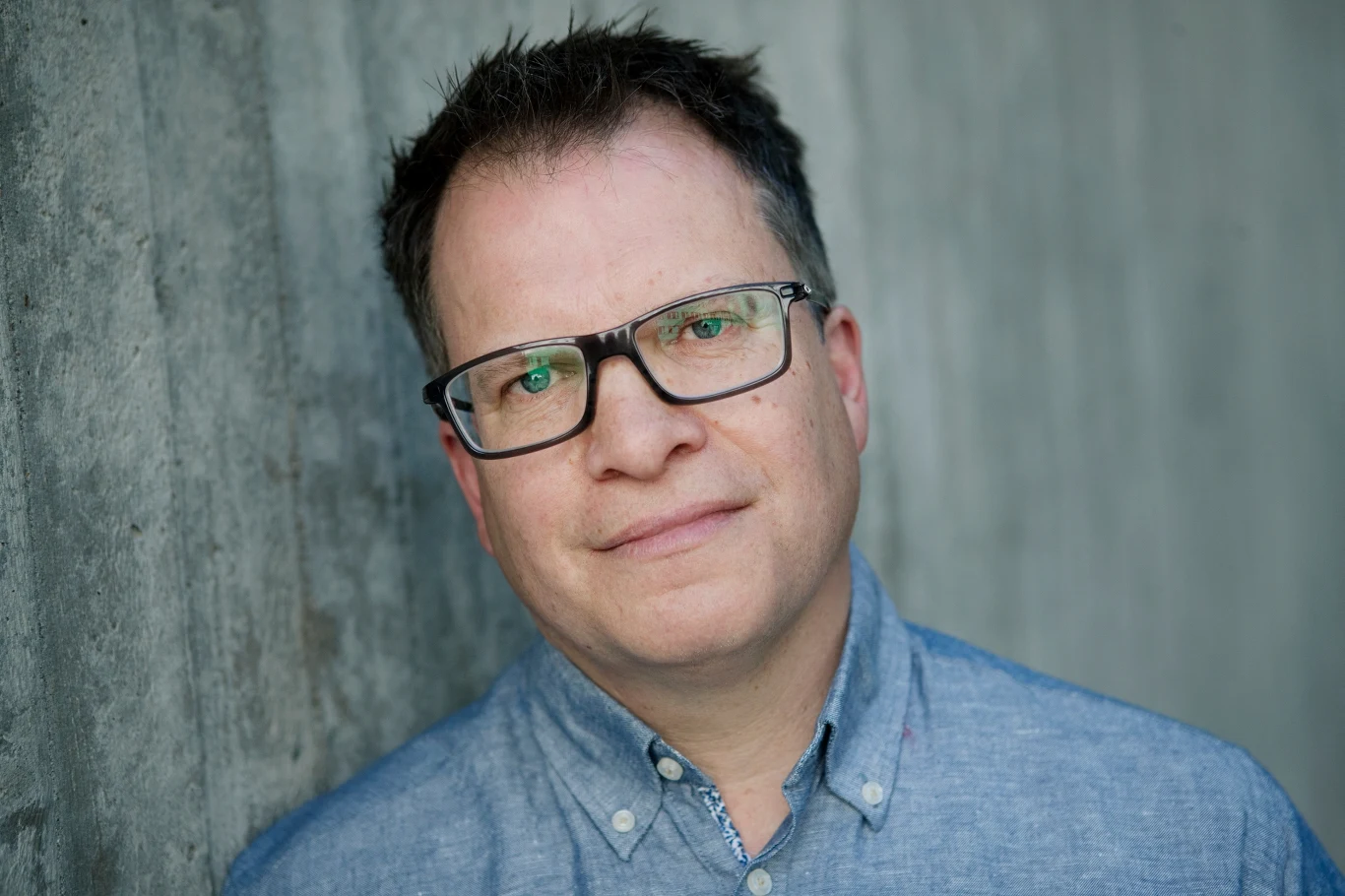 mężczyzna w okularach z krótkimi ciemnymi włosami, ubrany w jasnoniebieską koszulę, stoi przy betonowej ścianie i patrzy spokojnie w stronę obiektywu