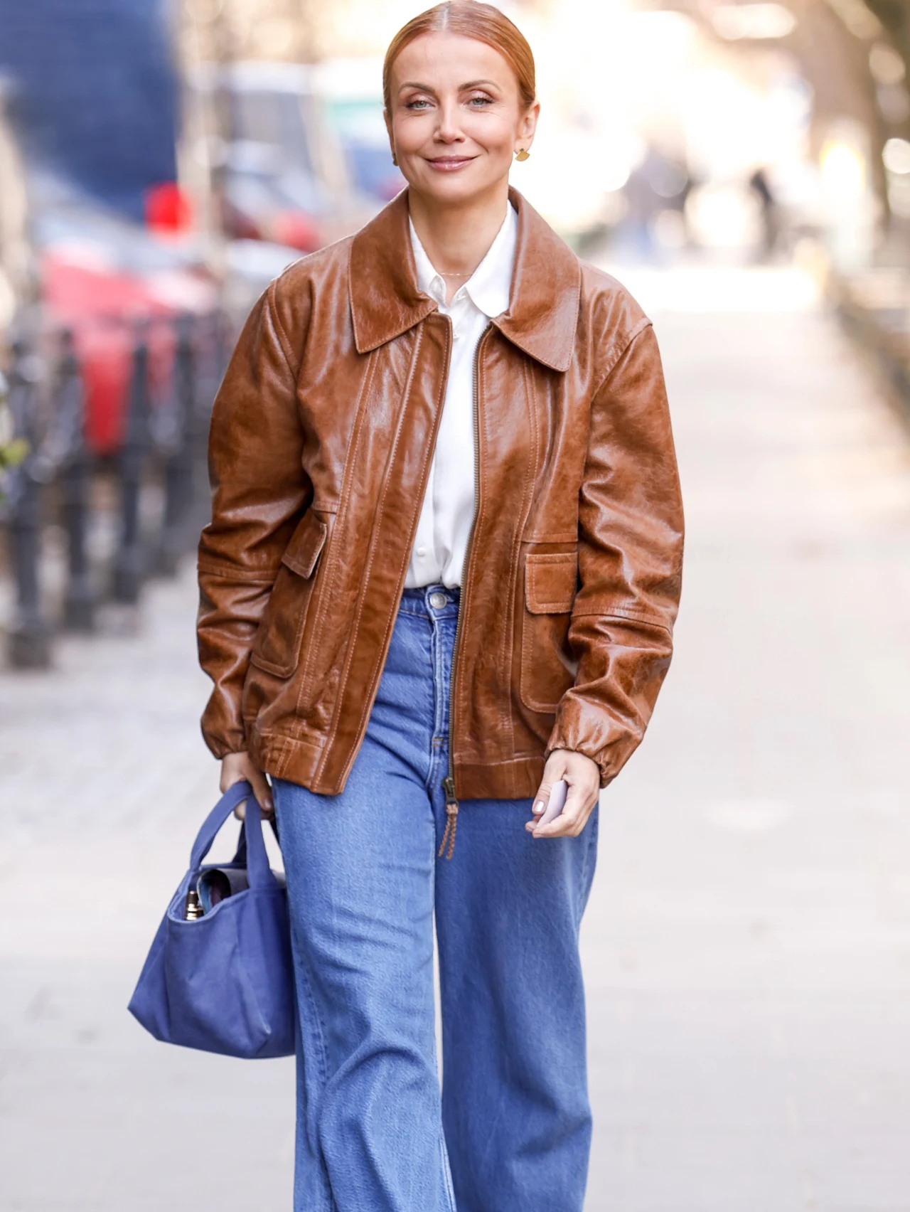 kobieta z jasnymi włosami spiętymi z tyłu idzie chodnikiem, ubrana w brązową skórzaną kurtkę, białą koszulę i szerokie niebieskie jeansy, trzymając w lewej ręce niebieską torebkę