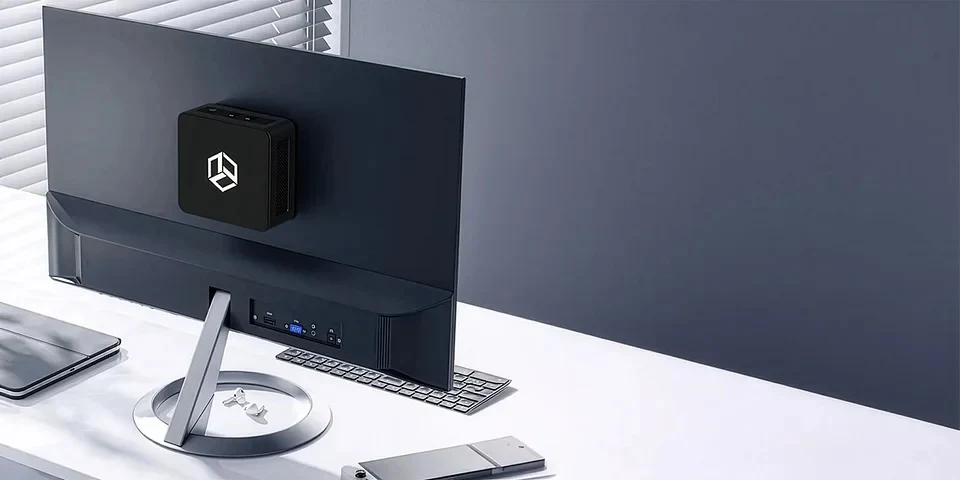 Nowoczesne stanowisko pracy z dużym monitorem komputerowym, do którego z tyłu przymocowany jest kompaktowy komputer mini PC z geometrycznym logo, klawiatura i smartfon leżące na białym biurku w minimalistycznym otoczeniu.