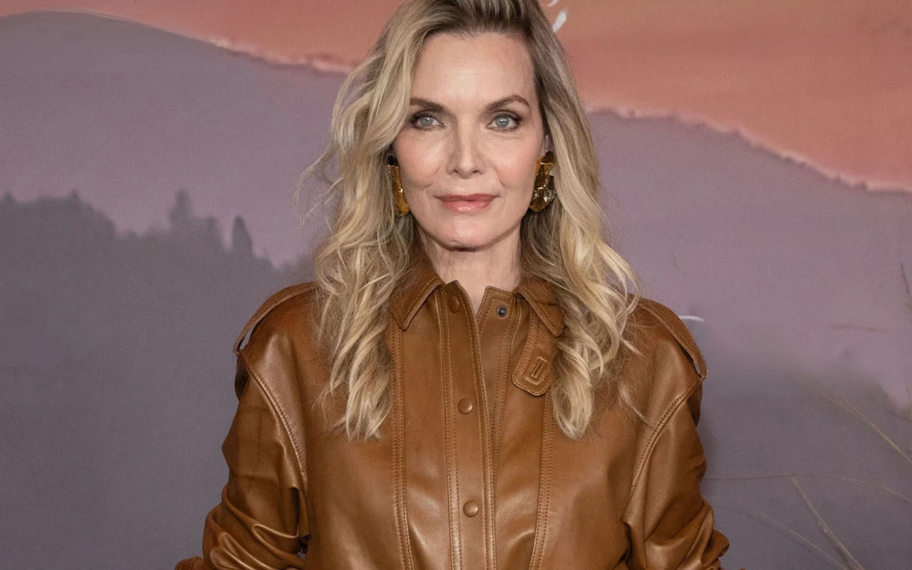 Kobieta o długich, falowanych blond włosach w skórzanej, brązowej kurtce, stojąca na tle rozmytego krajobrazu w odcieniach fioletu i różu.