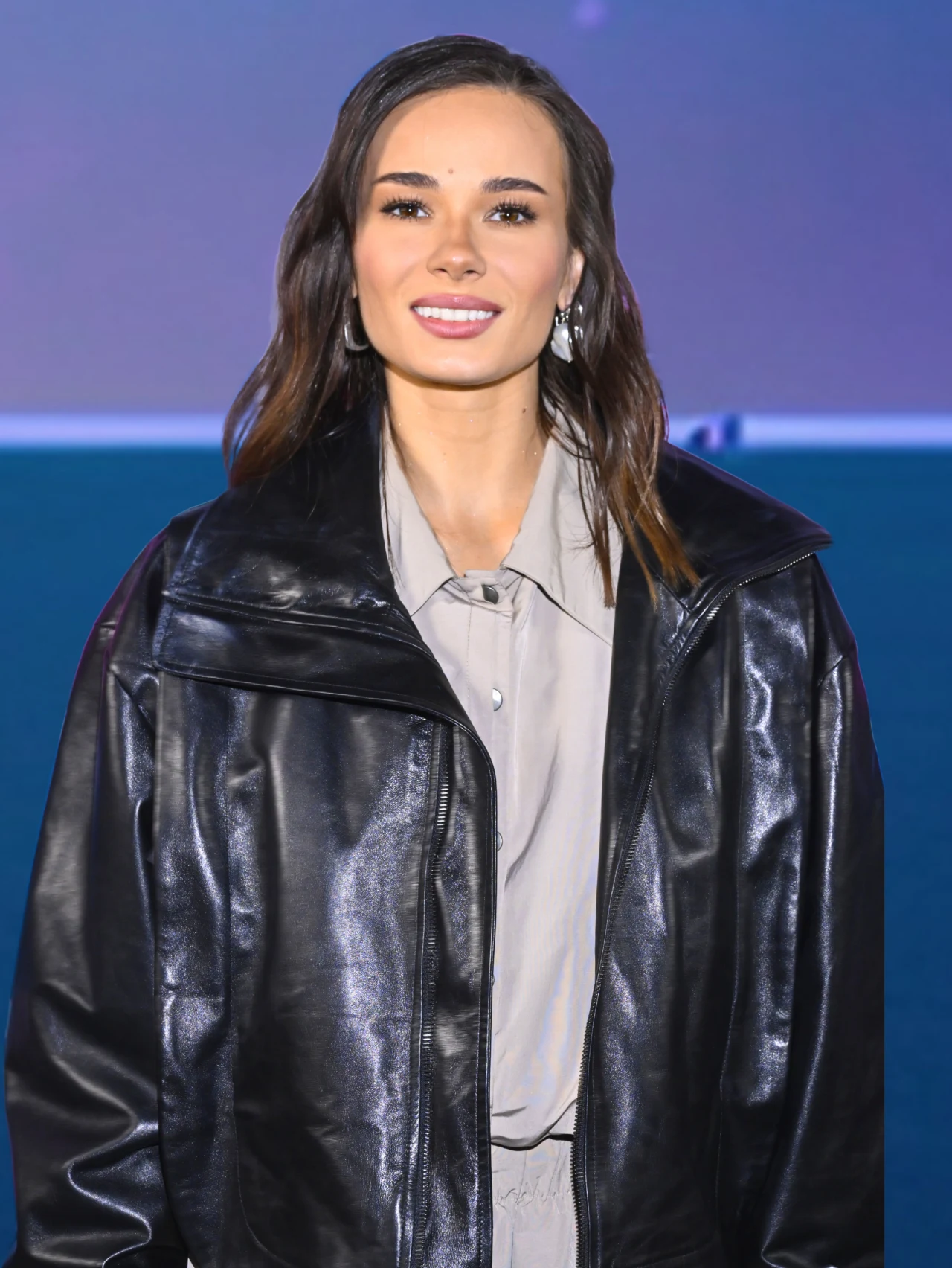 Uśmiechnięta kobieta z długimi, ciemnymi włosami, ubrana w czarną, skórzaną kurtkę i szarą koszulę, stoi na tle w niebieskich i fioletowych odcieniach.