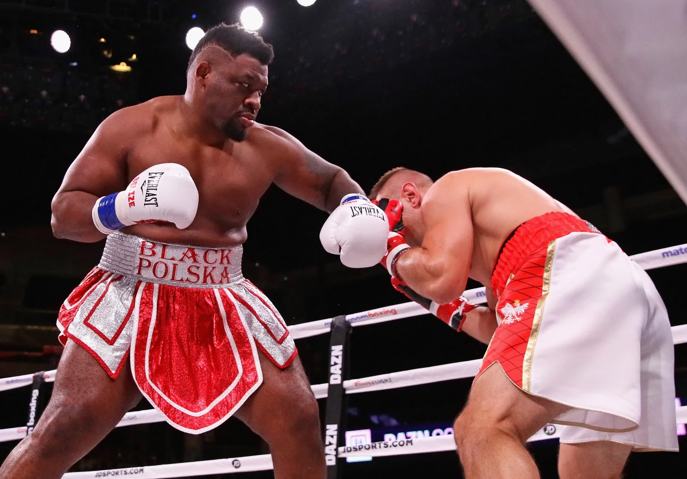 dwóch bokserów walczy na ringu, jeden z nich w czerwono-białych spodenkach z napisem „BLACK POLSKA” zadaje silny cios przeciwnikowi, który chroni głowę i jest zgarbiony w pozycji obronnej