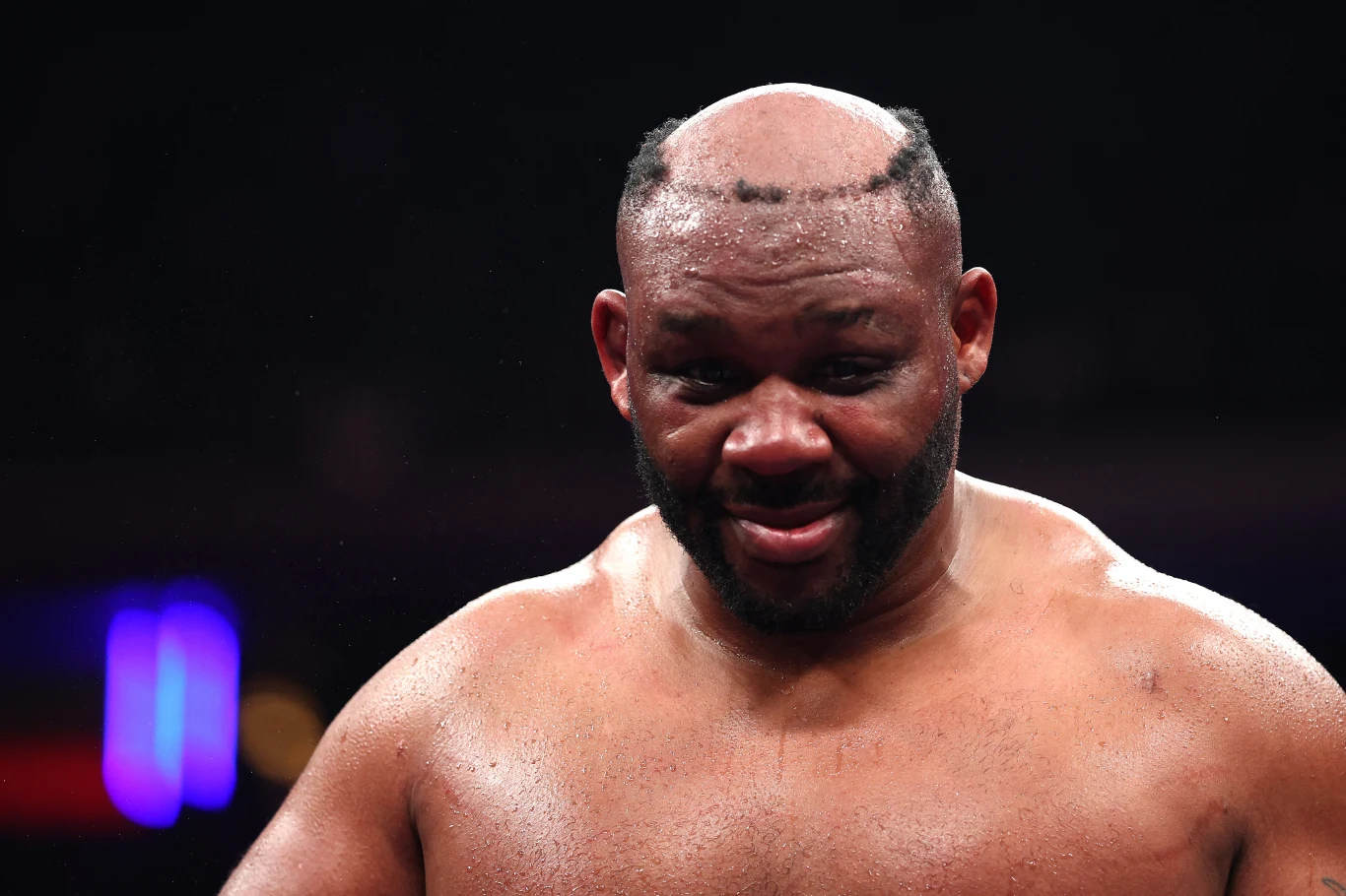 Mięśniowy mężczyzna z widoczną łysiną i bardzo nietypową fryzurą ograniczoną do wąskiego pasa włosów na obrzeżach głowy, stoi spocony na ringu bokserskim, z lekkim uśmiechem na twarzy.