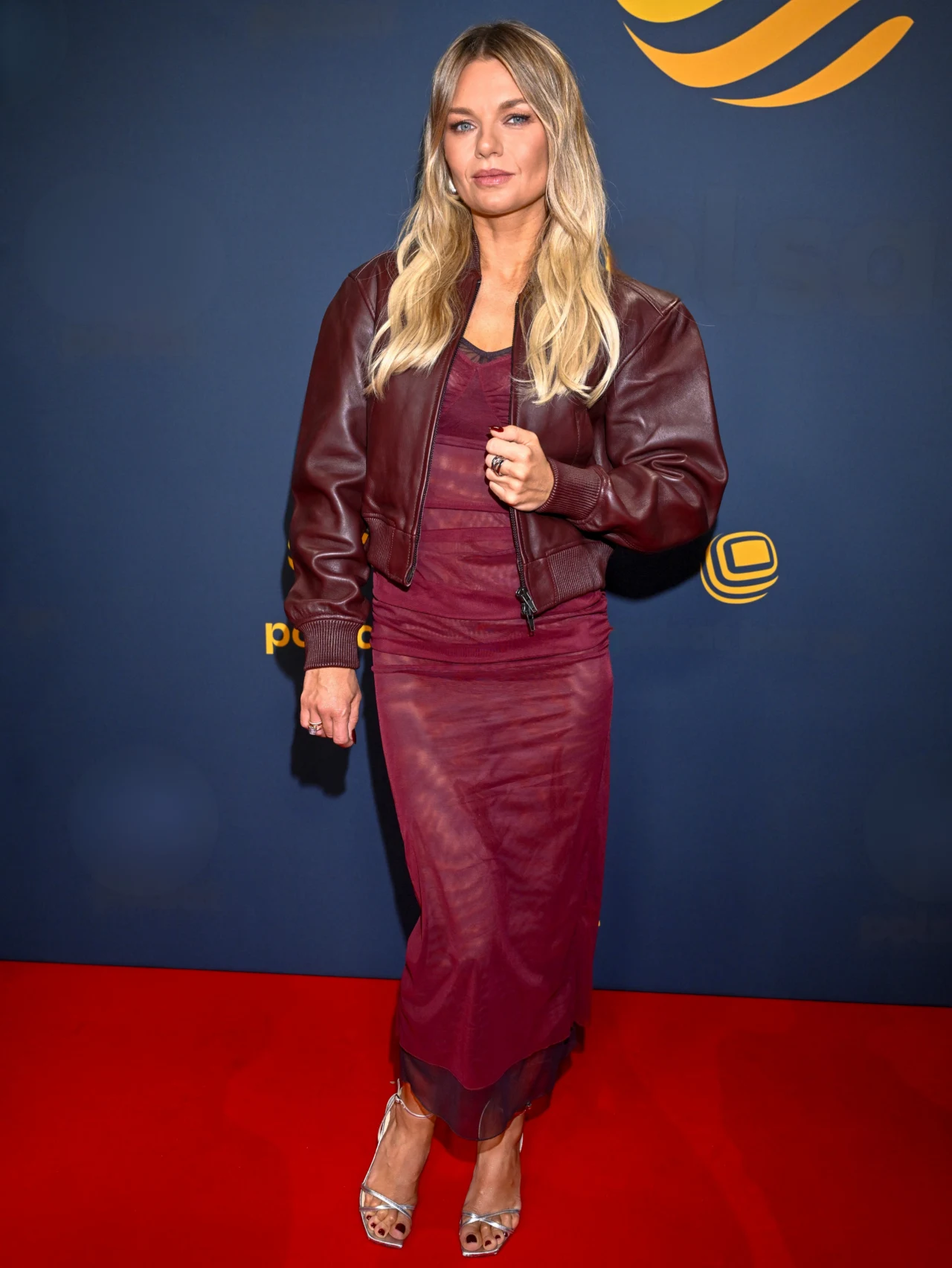 kobieta w bordowej sukience maxi i skórzanej kurtce stojąca na czerwonym dywanie, blond włosy rozpuszczone, jasne sandały na obcasie, granatowe tło z żółtym logotypem