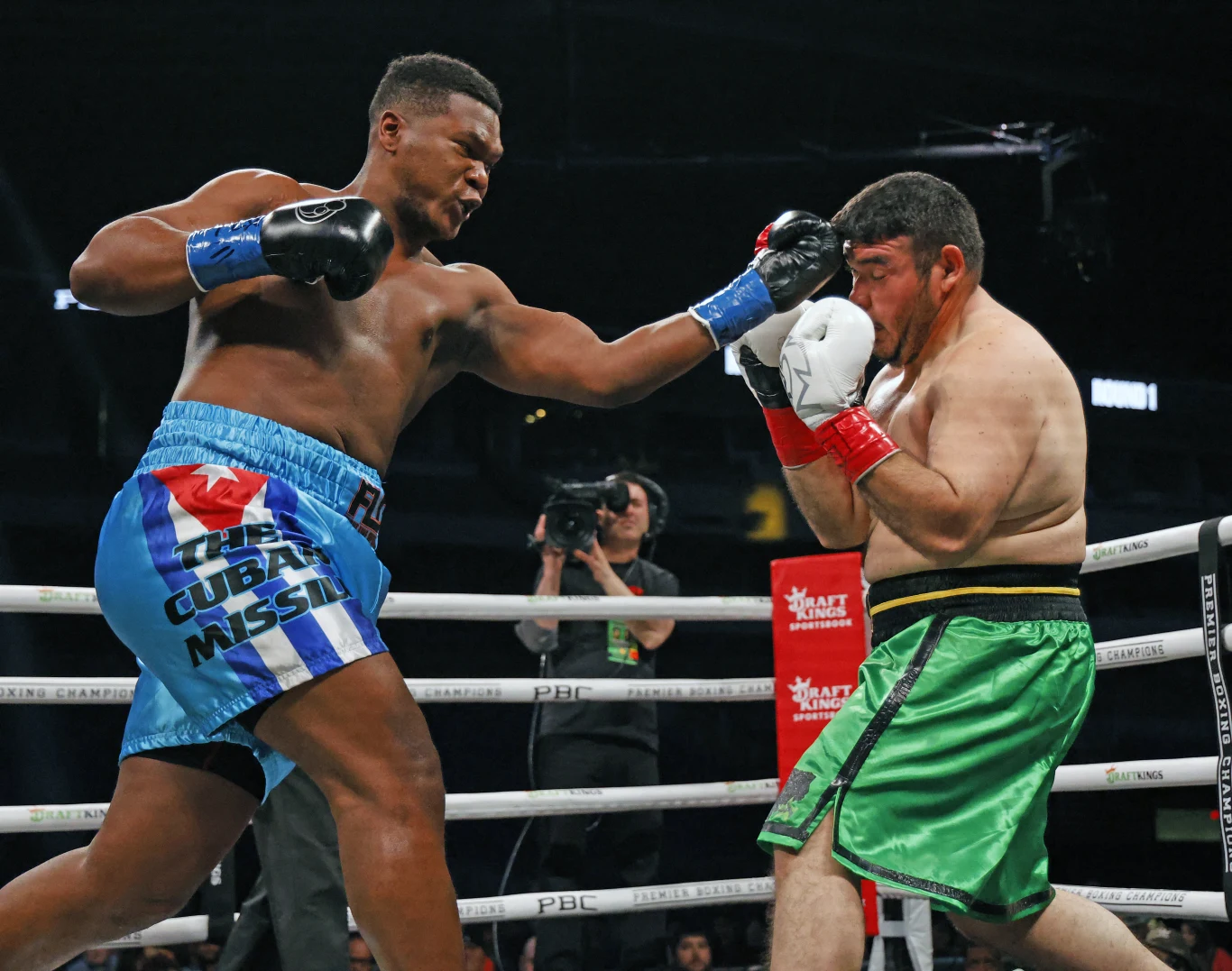 Dwóch bokserów walczących na ringu, jeden z nich w niebiesko-białych spodenkach zadaje mocny lewy cios przeciwnikowi w zielonych spodenkach, który próbuje zasłonić twarz rękawicami.