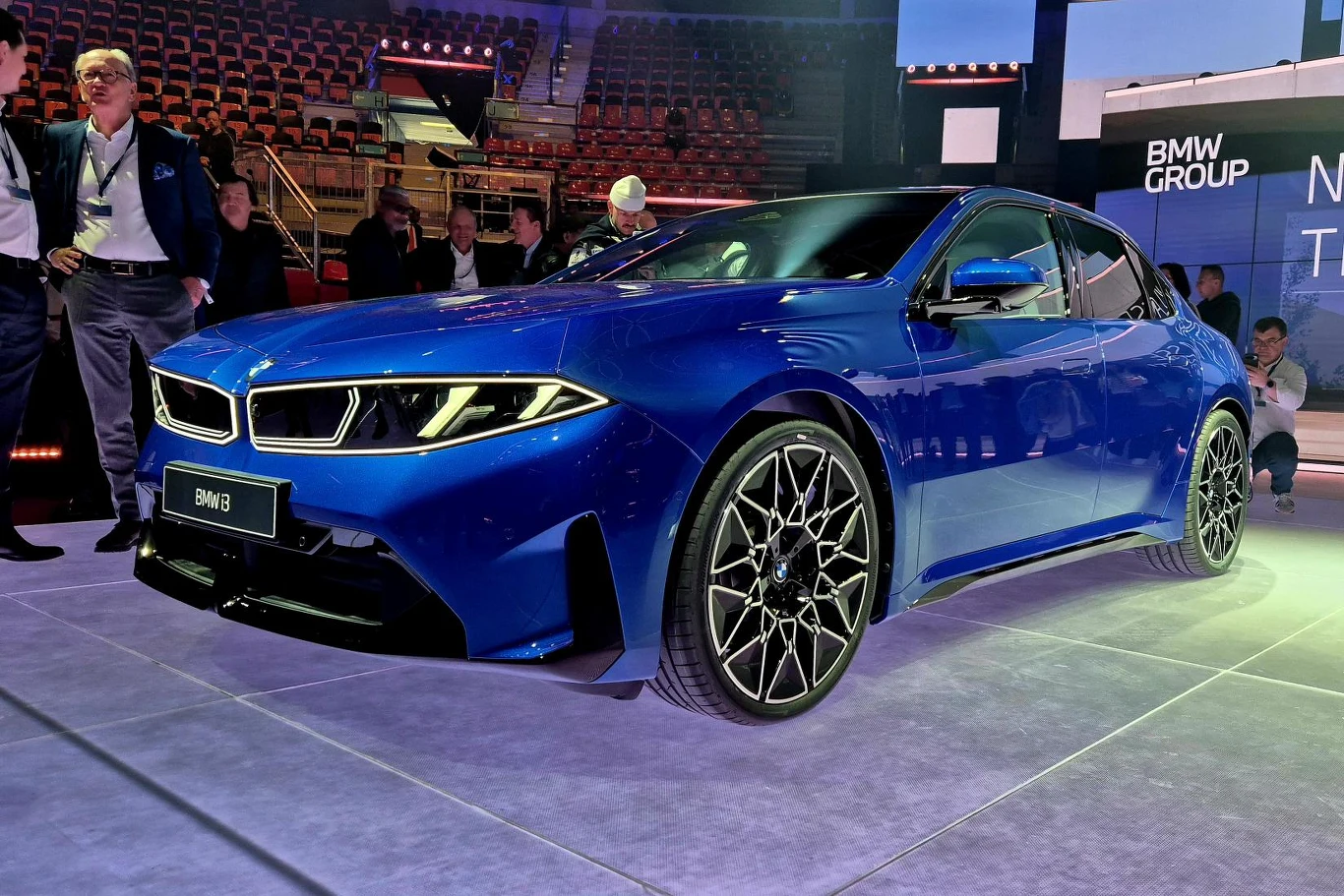 Nowoczesny, niebieski samochód BMW na ekspozycji, otoczony przez kilka osób obserwujących pojazd, reflektory LED o agresywnym kształcie i duże, stylowe felgi podkreślające sportowy charakter auta.