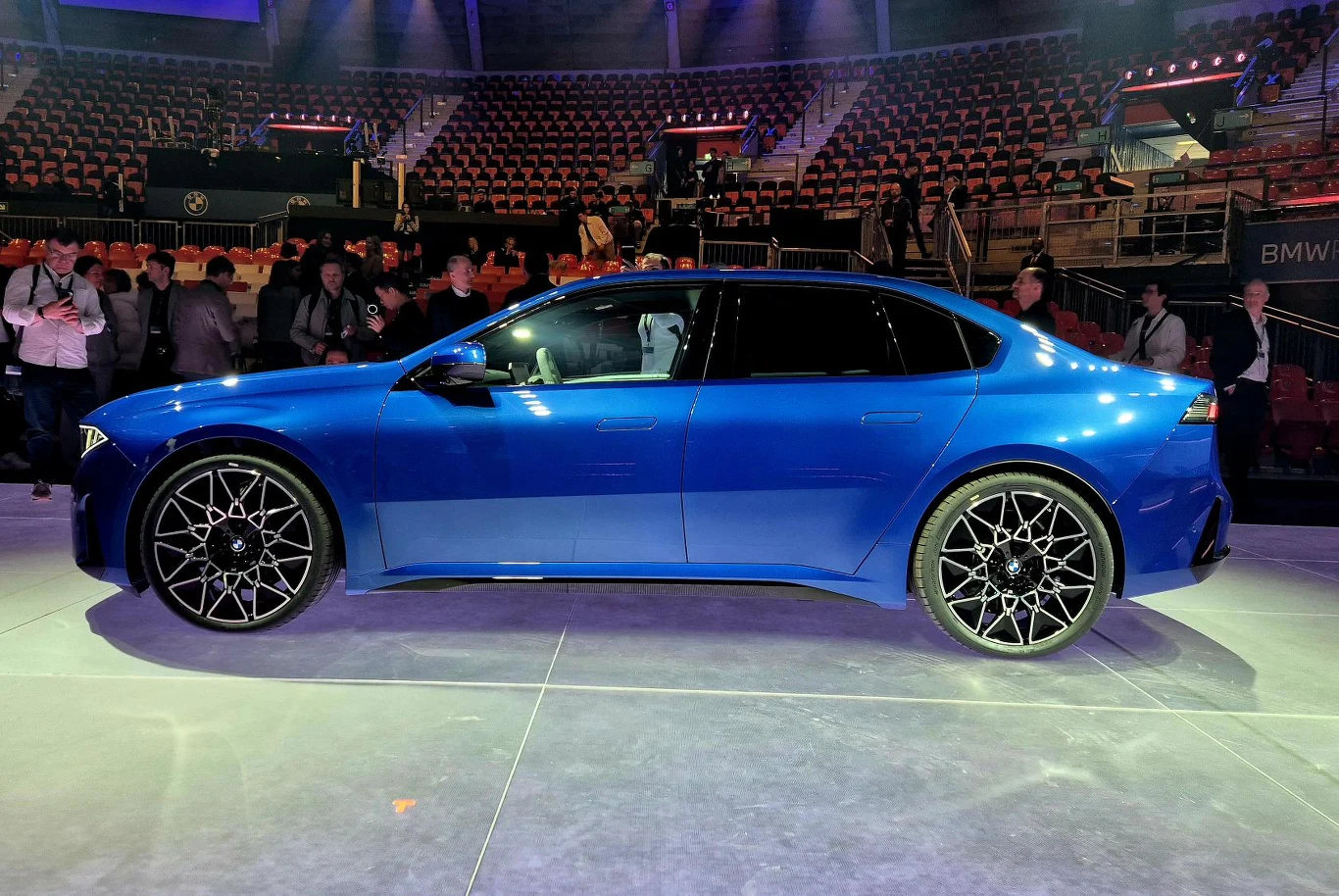 niebieski sportowy sedan BMW stoi na dobrze oświetlonej ekspozycji, wokół którego zgromadziła się grupa ludzi obserwujących samochód, na trybunach widać niewielu widzów