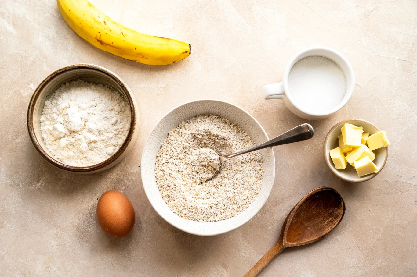 Składniki na ciasto lub deser: banan, mleko w białej filiżance, kostki masła w małej misce, jajko, miska z mąką, miska z wymieszanymi składnikami i łyżką oraz drewniana łyżka na jasnym tle.
