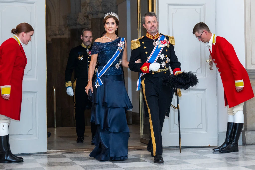 elegancka para w strojach galowych wychodzi przez otwarte drzwi, mężczyzna w mundurze z licznymi orderami i kobieta w granatowej sukni z diademem na głowie, oboje otoczeni przez dworzan w czerwonych mundurach składających ukłon