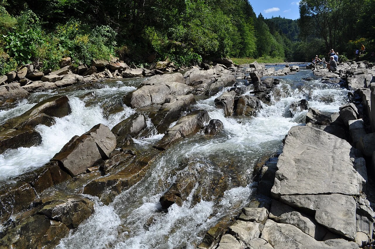 Górski potok płynący przez kamieniste koryto, otoczony zielonym lasem; w tle widać kilka osób spacerujących po brzegu.