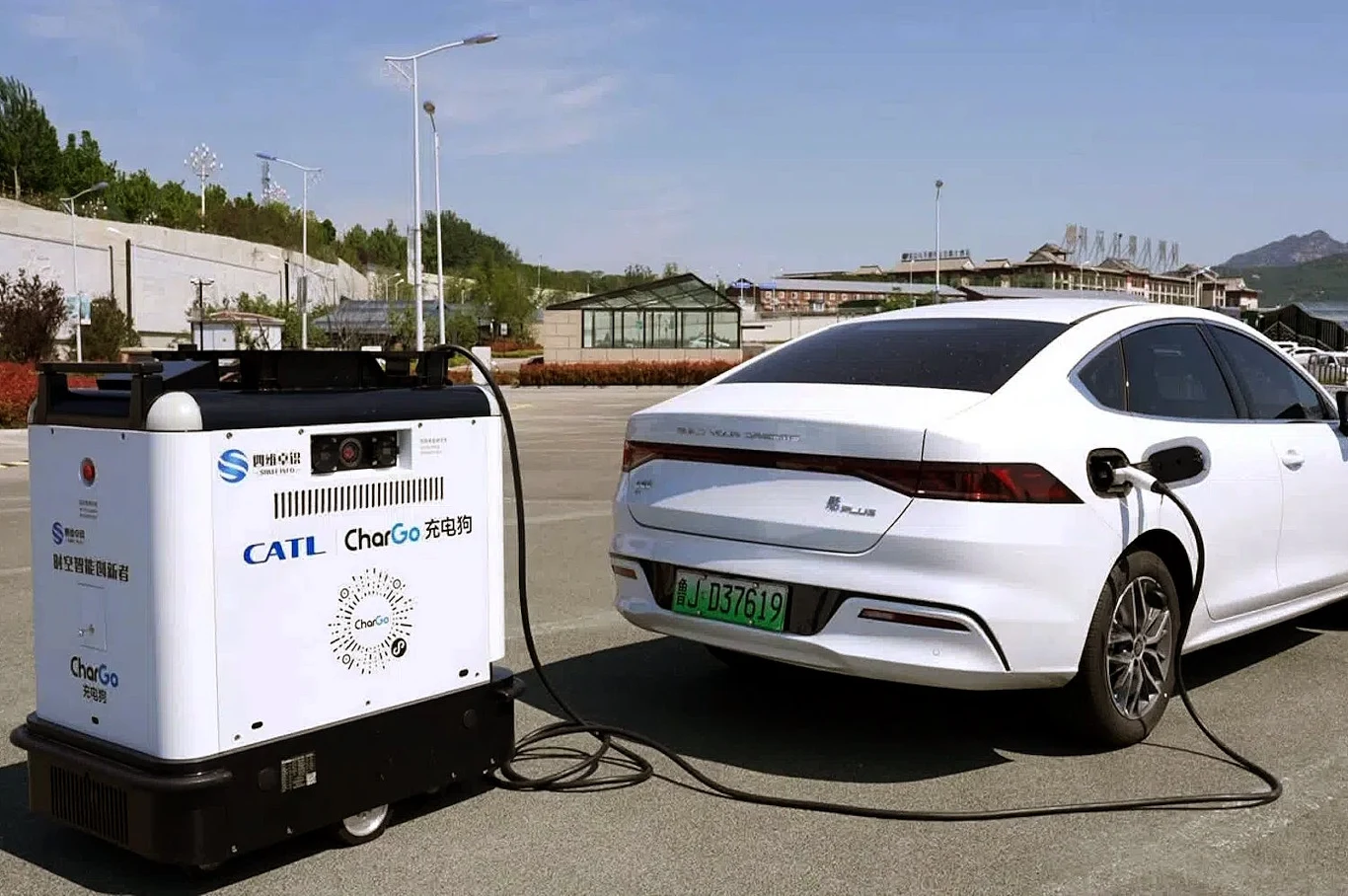biały elektryczny samochód podłączony do przenośnej ładowarki stojącej na parkingu; kabel łączy urządzenie z gniazdem ładowania auta, a wokół znajduje się przestrzeń miejska z niską zabudową i roślinnością w tle