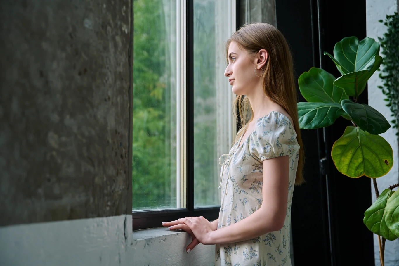 Młoda kobieta o jasnych włosach w zwiewnej sukience stoi przy dużym oknie, patrząc zamyślona na zewnątrz; obok niej zielona roślina doniczkowa.