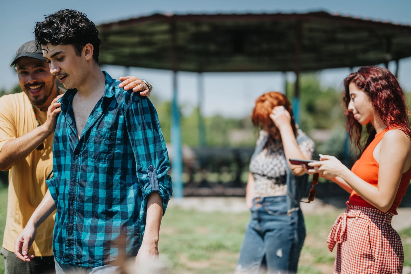 Czworo młodych ludzi spędza czas na świeżym powietrzu, jedna osoba uśmiecha się i kładzie rękę na ramieniu drugiej, obok kobieta sprawdza coś w telefonie, a w tle kolejna rozmawia przez telefon.