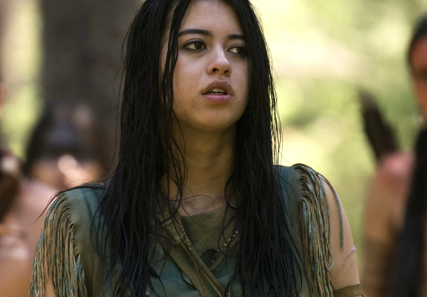 "Predator: Łowca" Młoda kobieta z długimi ciemnymi włosami w zielonej tunice; w tle jasny las, kadr z filmu "Predator: Łowca".