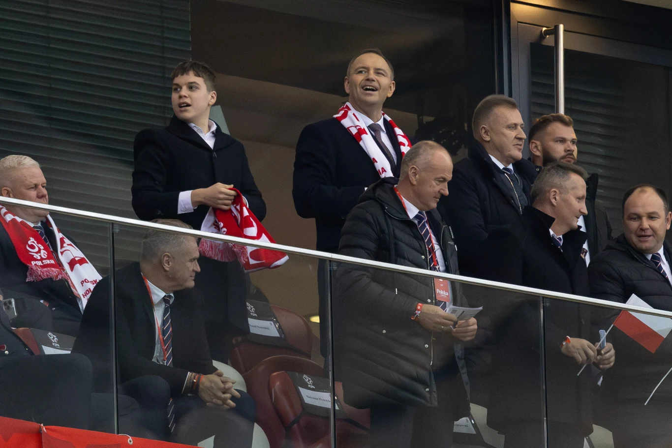 Grupa elegancko ubranych mężczyzn obserwuje mecz z loży stadionu, część z nich trzyma biało-czerwone szaliki i flagę, wszyscy skupieni na wydarzeniach na boisku.