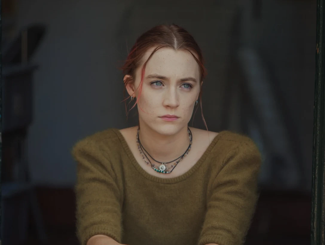 Kadr z filmu "Lady Bird" Młoda kobieta o jasnych włosach spiętych w luźny kok, ubrana w zielony sweter, patrząca zamyślonym wzrokiem w dal
