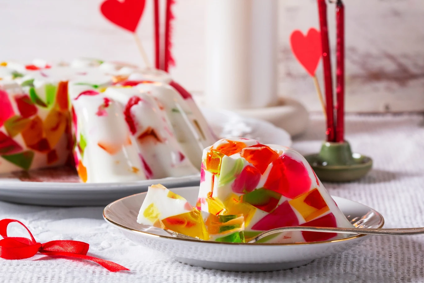 Kolorowa galaretka zatopiona w białej masie śmietankowej pokrojona na kawałki na talerzu, w tle czerwone dekoracje w kształcie serca i zapalone świece.