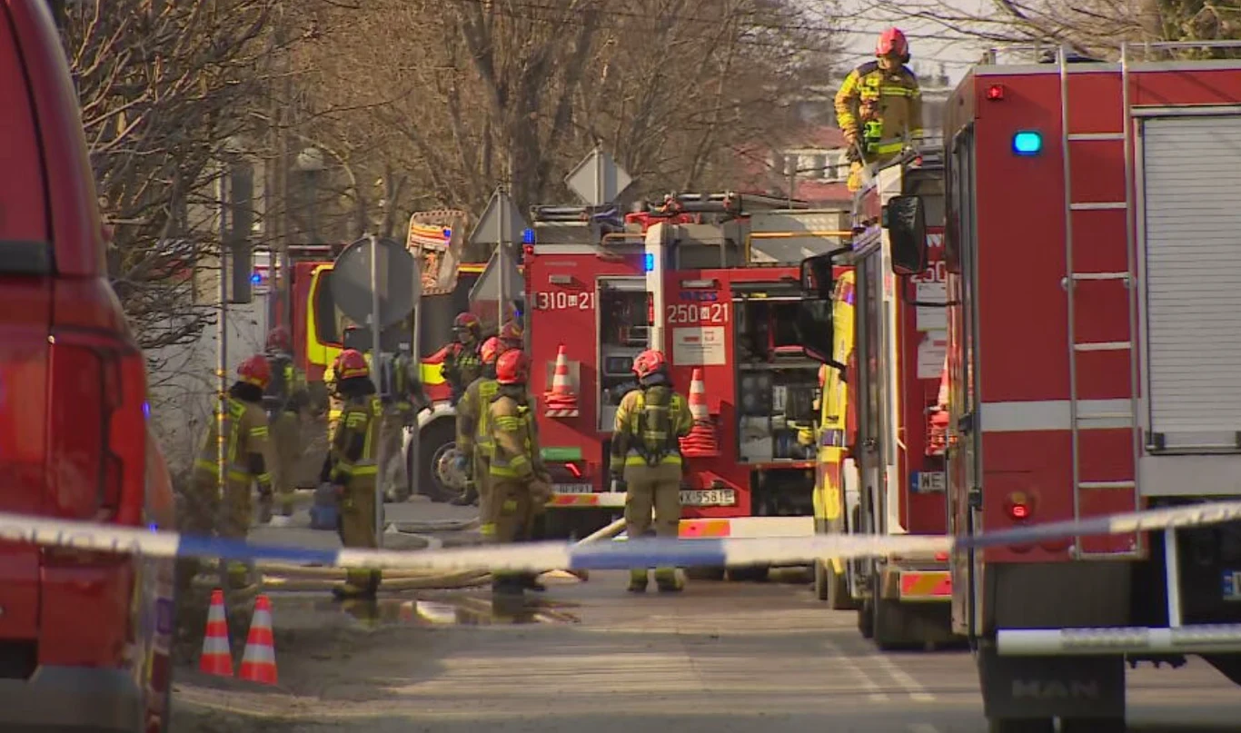 Strażacy w specjalistycznych ubraniach prowadzą akcję ratowniczo-gaśniczą wokół kilku wozów strażackich ustawionych na miejskiej ulicy, miejsce zabezpieczone taśmą i pachołkami, w tle drzewa i oznakowanie drogowe.